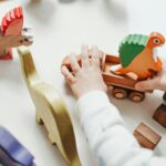 Child's hands interact with vibrant wooden toy dinosaur set, creating playful scene on tabletop.