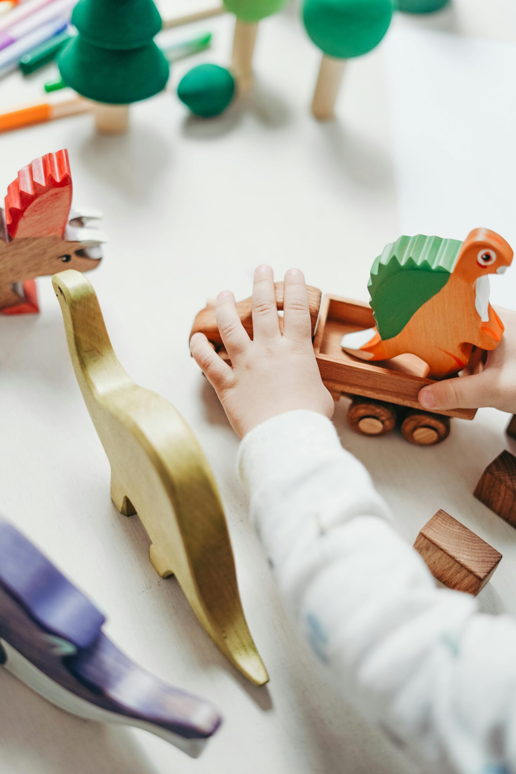 Child's hands interact with vibrant wooden toy dinosaur set, creating playful scene on tabletop.